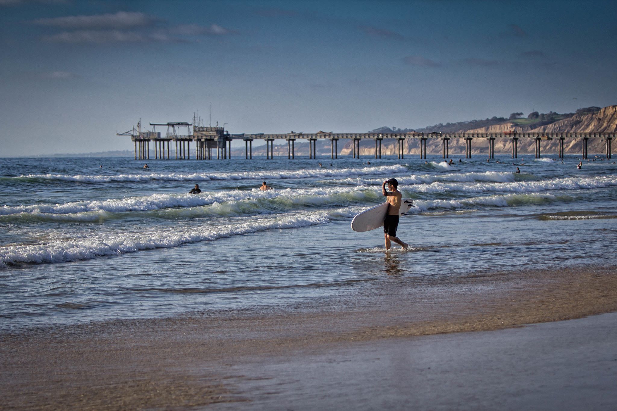 surfer walking into surf; distant pier extends from bluff into ocean